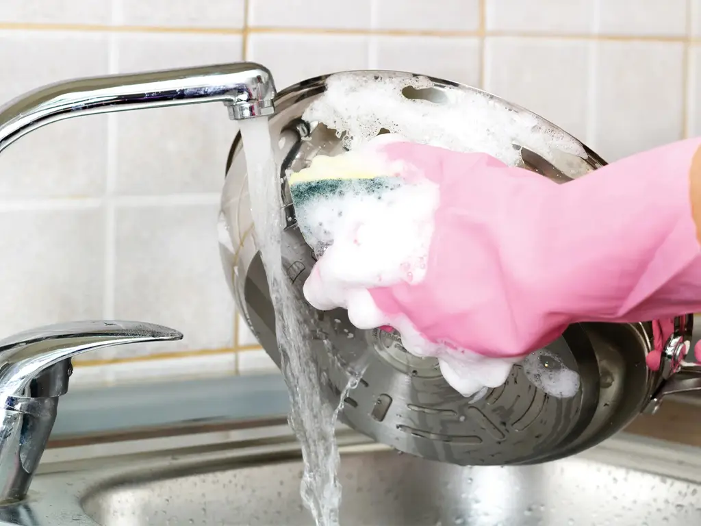 Personne avec un gant rose lavant une casserole à la main sous l'eau du robinet - réparation lave-vaisselle Genève, Suisse Dépannage